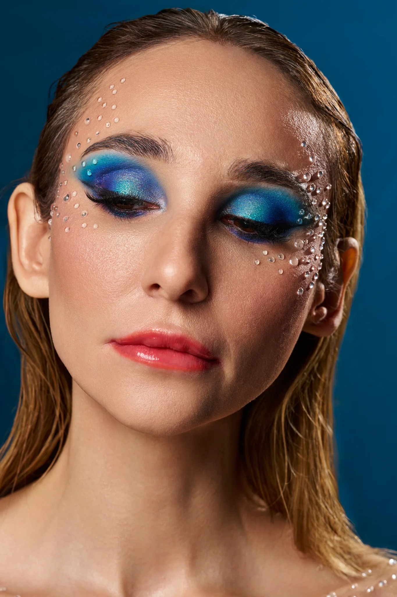 Studio beauty portrait with deep blue eyeshadow and crystal face details, wet hair, eyes closed, blue backdrop