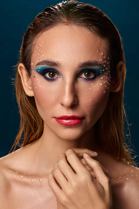 Studio beauty portrait with blue editorial eye makeup, crystal face details, hand at collarbone, dark blue background