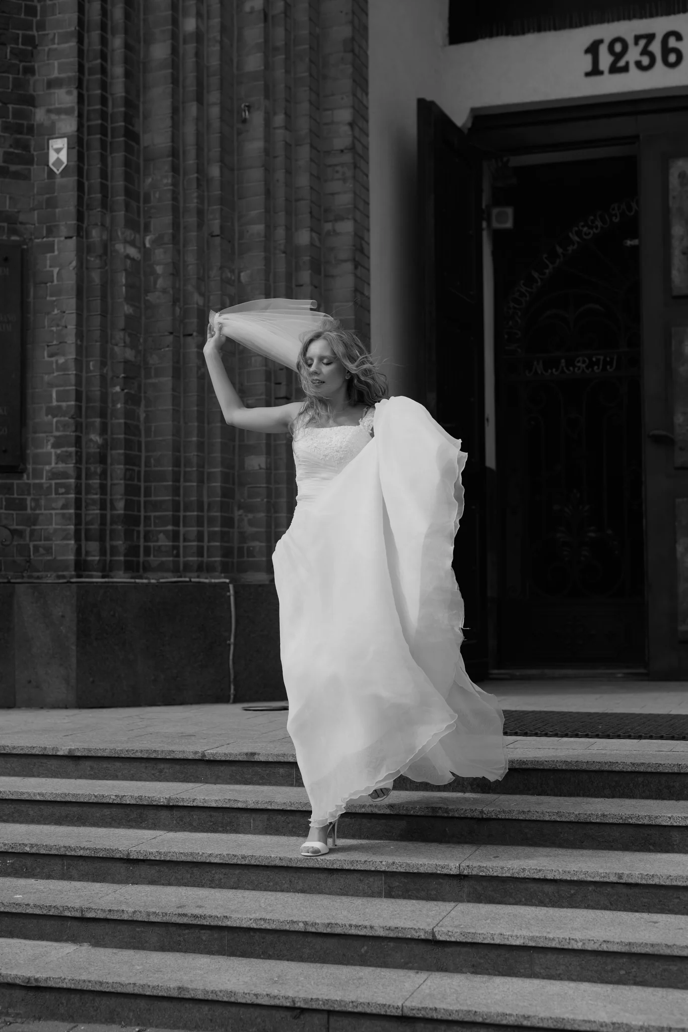 Woman in wedding dress running down church steps, veil raised above her head, black and white outdoor portrait, Poland.