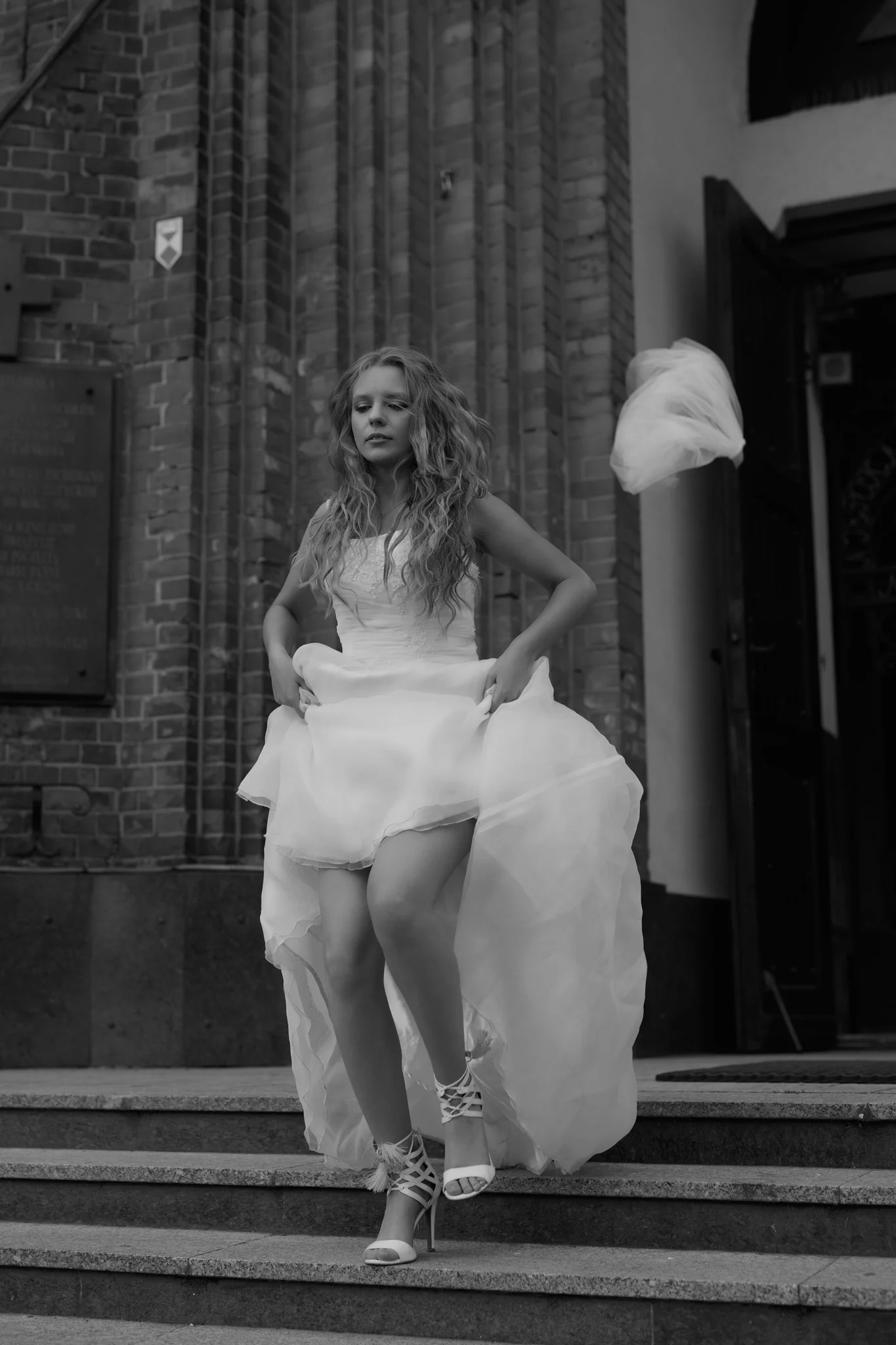 Woman in wedding dress standing on church steps, dress caught mid-motion, veil flying in the background, hands on hips, black and white outdoor portrait, Poland.