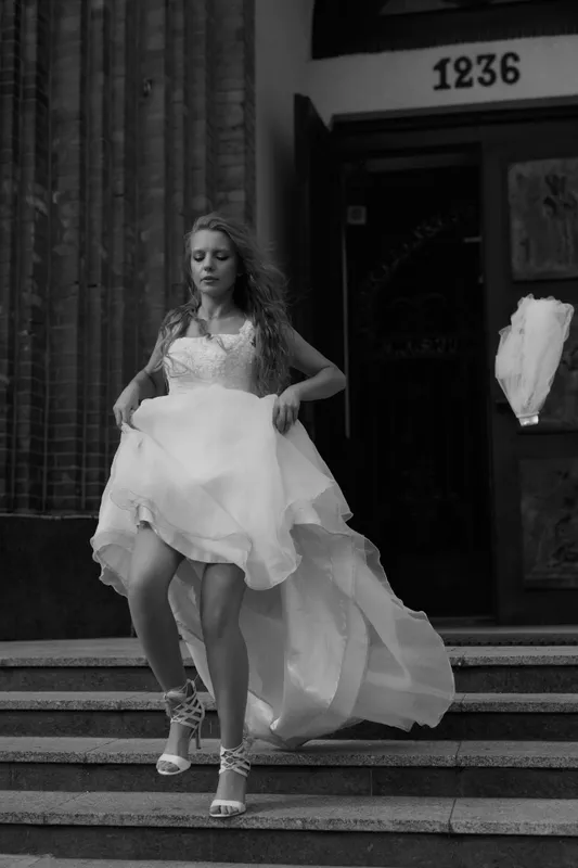 Woman in wedding dress on church steps, dress lifted mid-motion, veil floating in the background, black and white outdoor portrait.