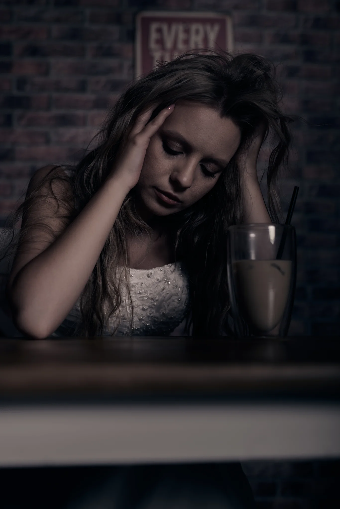 Woman in wedding dress at a café table, head bowed, both hands at her face, exhausted pose.