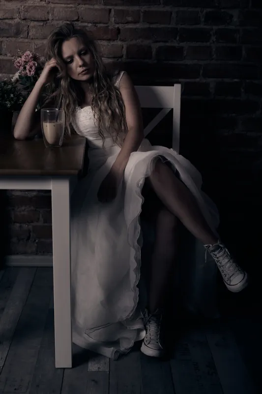 Woman in wedding dress sitting on a chair at a café table with brick wall background, wearing Converse sneakers, flowers in the background.
