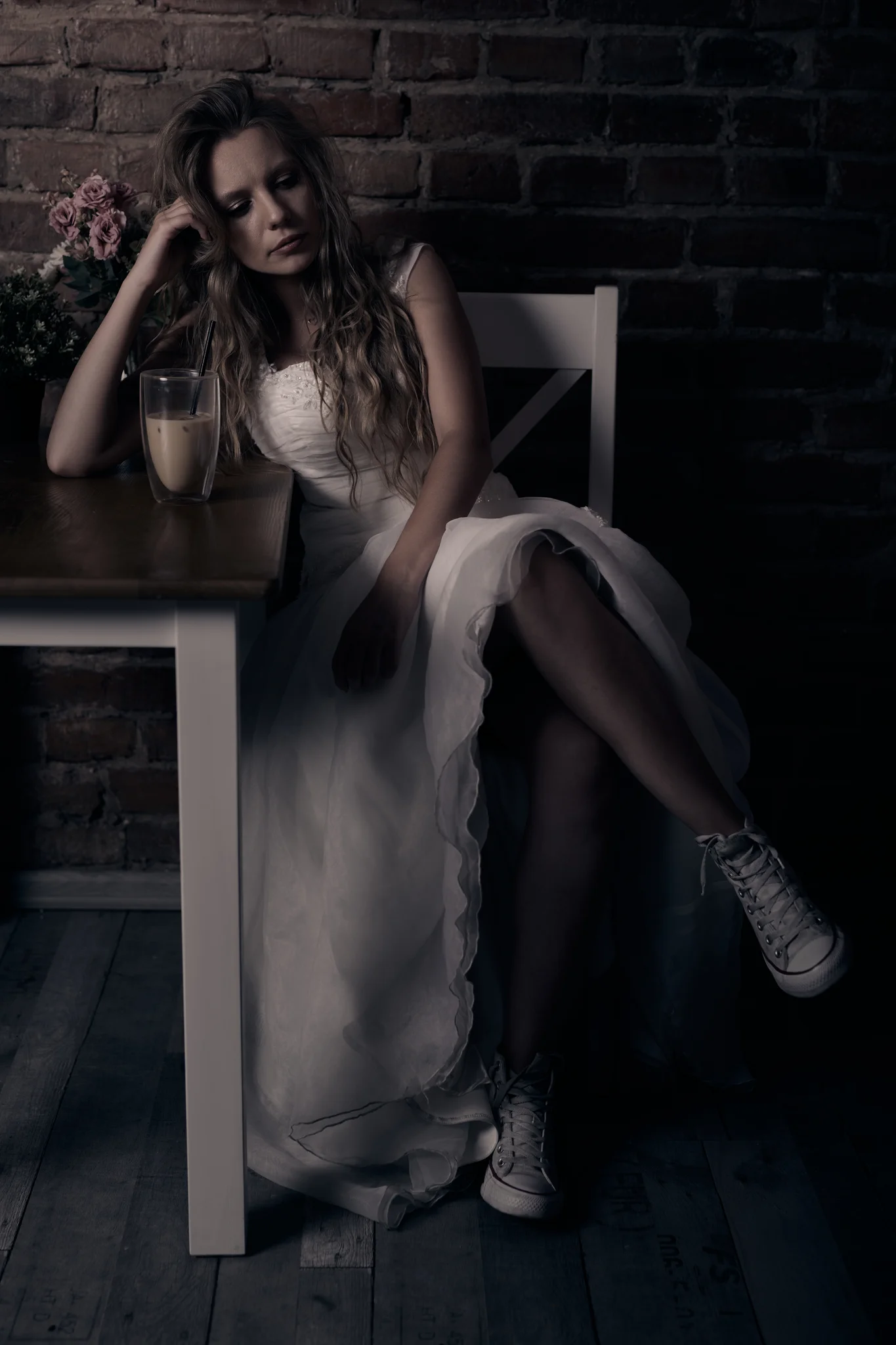 Woman in wedding dress sitting on a chair at a café table with brick wall background, wearing Converse sneakers, flowers in the background.